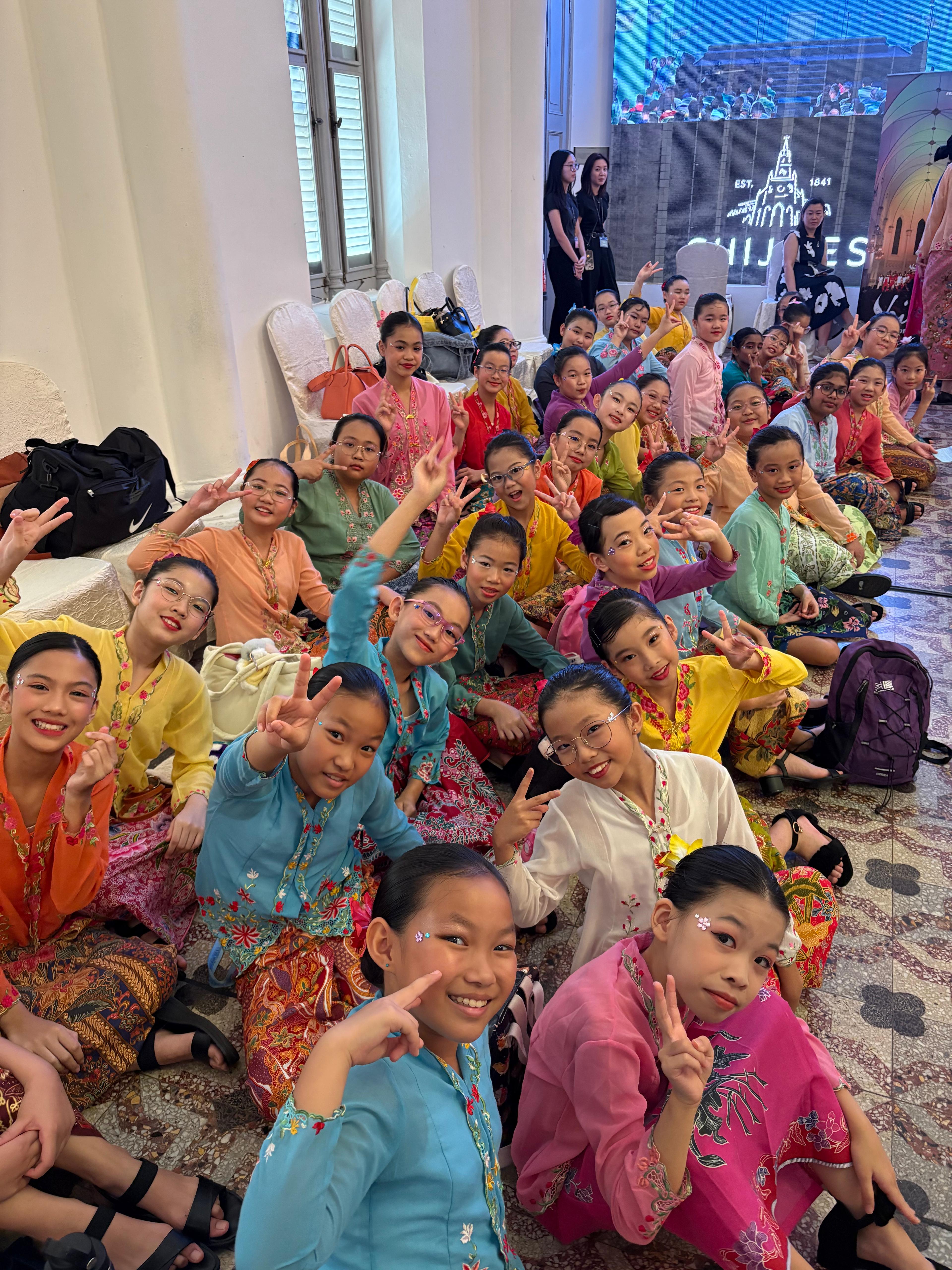 A large group of young girls in colorful traditional Southeast Asian costumes sit together smiling and making peace signs.